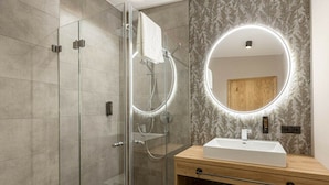 Un bany modern amb dutxa de vidre, un tocador de fusta amb un lavabo blanc i un mirall rodó amb il·luminació integrada.