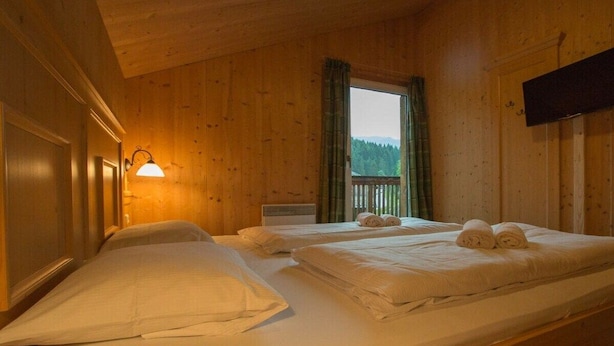 Una habitación con dos camas, una ventana con cortinas, un televisor montado en la pared y techo de madera.