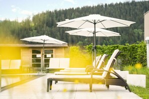 A poolside area with lounge chairs and umbrellas, a wooden building in the background, and a forested hillside. - Apartment in Radstadt with swimming pool (Radstadt)