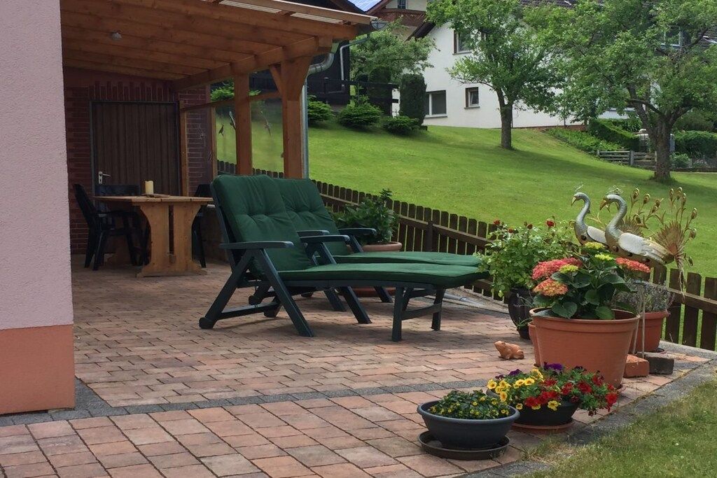 Un patio cubierto con una silla de descanso verde, una mesa y plantas en macetas.