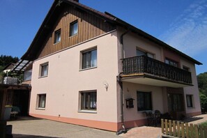 A two-story building with a balcony, a covered outdoor seating area, and a visible garage entrance. - Holiday apartment Brigitte DG (Flörsbachtal)