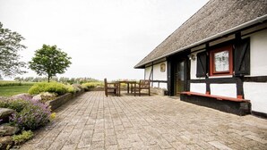 A building with a stone-paved patio, wooden furniture, and a view of a green landscape. - Nydamsgaard "hus mælkebøtte" (Faaborg \/ Bojden)