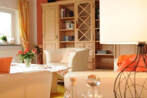 Ein gemütliches Zimmer mit einem weißen Sessel, einem kleinen Tisch mit Blumenvase und einer Bücherwand voller Bücher.