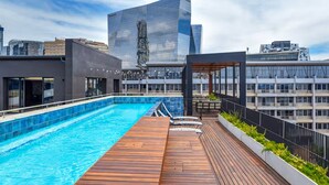 Piscina en la azotea con plataforma de madera, rodeada de edificios modernos.