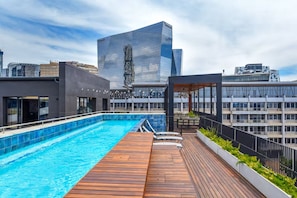 Piscina en la azotea con plataforma de madera, rodeada de edificios modernos.