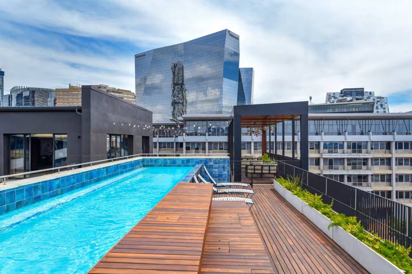 Una piscina en la azotea con un deck de madera y un edificio de vidrio al fondo.