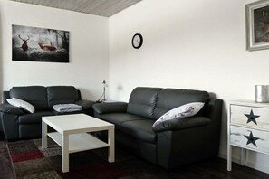 Un salon avec deux canapés noirs, une table basse blanche, une horloge au mur et une table de chevet ornée d’un motif en étoile.