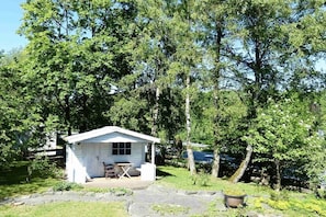 Une petite cabane blanche avec un porche et une table, entourée d’arbres et bordée d’un chemin de pierres.