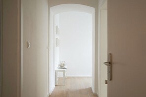 Ein Flur mit einer weißen Tür rechts, einem kleinen Tisch mit einer Vase und einem Buch sowie einem Lichtschalter an der Wand.