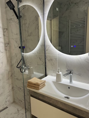 Un baño con ducha envuelta en vidrio, un lavabo blanco con grifo moderno y un espejo grande con iluminación circular.