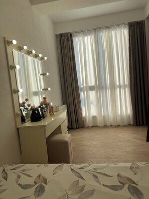 Habitación de hotel con tocador, espejo con luces, planta en maceta y ventana con cortinas.