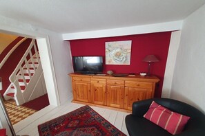 A living room with a television on a wooden cabinet, a red wall, a black chair with a red pillow, and a patterned rug. - Holiday home with 2 bedrooms (Bodenwerder)