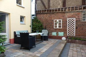 Een terras met buitenmeubilair, een bakstenen muur en een raam met een rankenmotief.