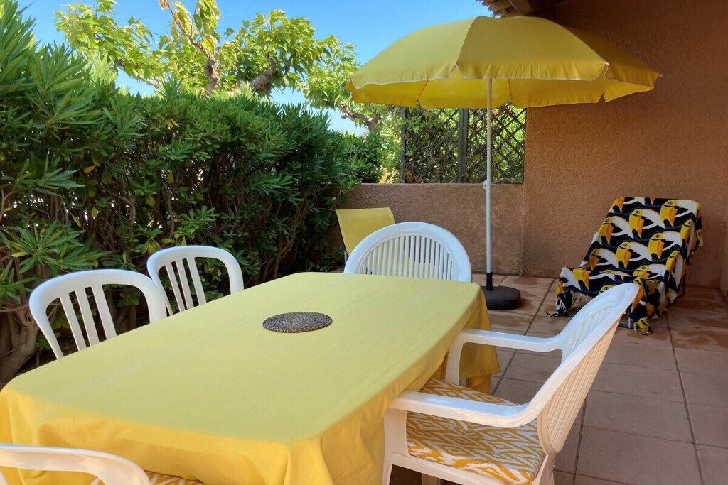 Une terrasse extérieure avec une table jaune et des chaises blanches, un parasol jaune et un canapé à motifs.