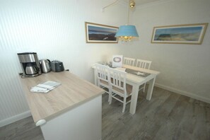A compact kitchenette with a coffee maker, toaster, and kettle. There is a small dining table with chairs and a wall-mounted lamp. Two framed pictures are hung above the table. - Holiday apartment in Sylt with 1 bedroom (Wenningstedt)