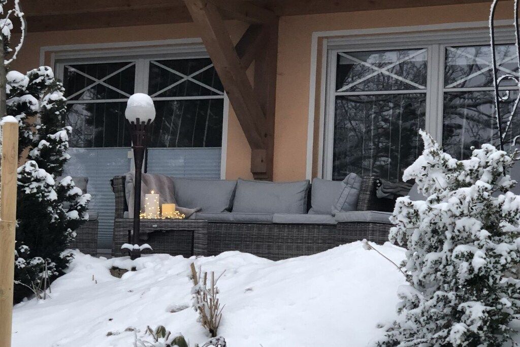 Un espace extérieur aménagé avec des sièges, recouvert de neige, comprenant un canapé en osier et une table illuminée, situé devant un bâtiment doté de grandes fenêtres.
