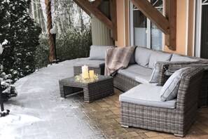 A covered outdoor seating area with a wicker sofa and a small table, surrounded by snow-covered ground and trees. - Holiday apartment in the Harz Mountains with 1 bedroom (Blankenburg)