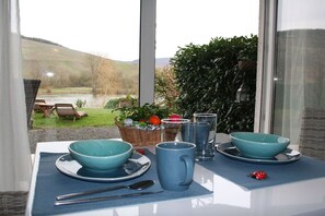 Une table à manger avec de la vaisselle bleue, dressée pour deux personnes, un panier de fruits et une vue sur une rivière et des collines.