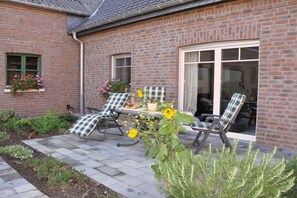 Een bakstenen huis met een terras, twee stoelen, een tafel met bloemen en een raam met bloembak.