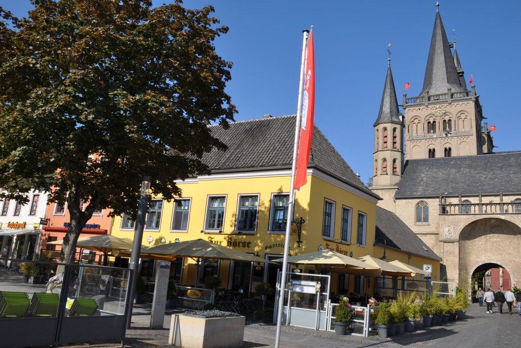 Een geel gebouw met terras, een vlaggenmast en een kerk met een toren op de achtergrond.
