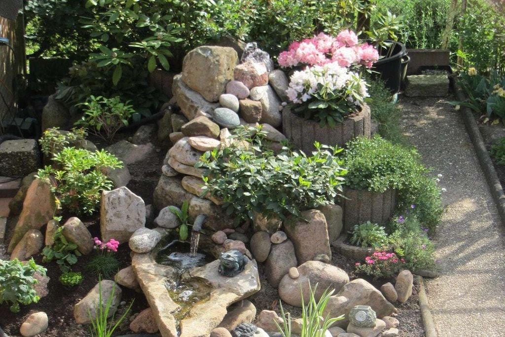 Ein Garten mit einem kleinen Wasserspiel, umgeben von Steinen und Pflanzen.