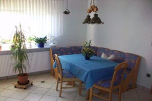 Uma área de jantar com mesa, cadeiras, sofá, plantas em vasos e luminárias pendentes.