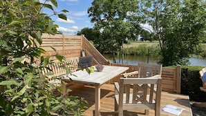 An outdoor wooden deck with a table and chairs, overlooking a body of water and surrounded by greenery. - Holiday cottage "Jantje" (Bunde)