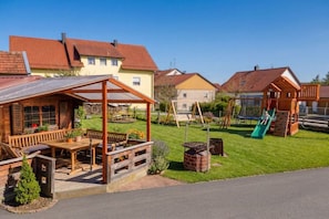 A wooden outdoor seating area with a table and benches, a brick fire pit, and a children's play structure with a slide. - at the Stark holiday farm (Kelheim)