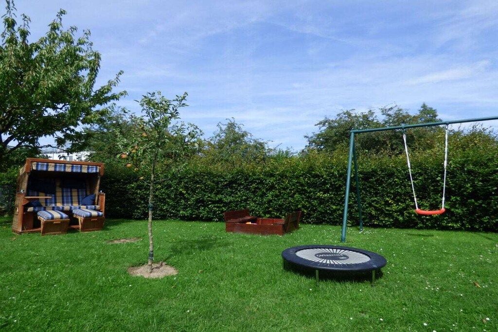 Un patio trasero con un trampolín, un conjunto de columpios y una casita de juegos de madera con asientos.