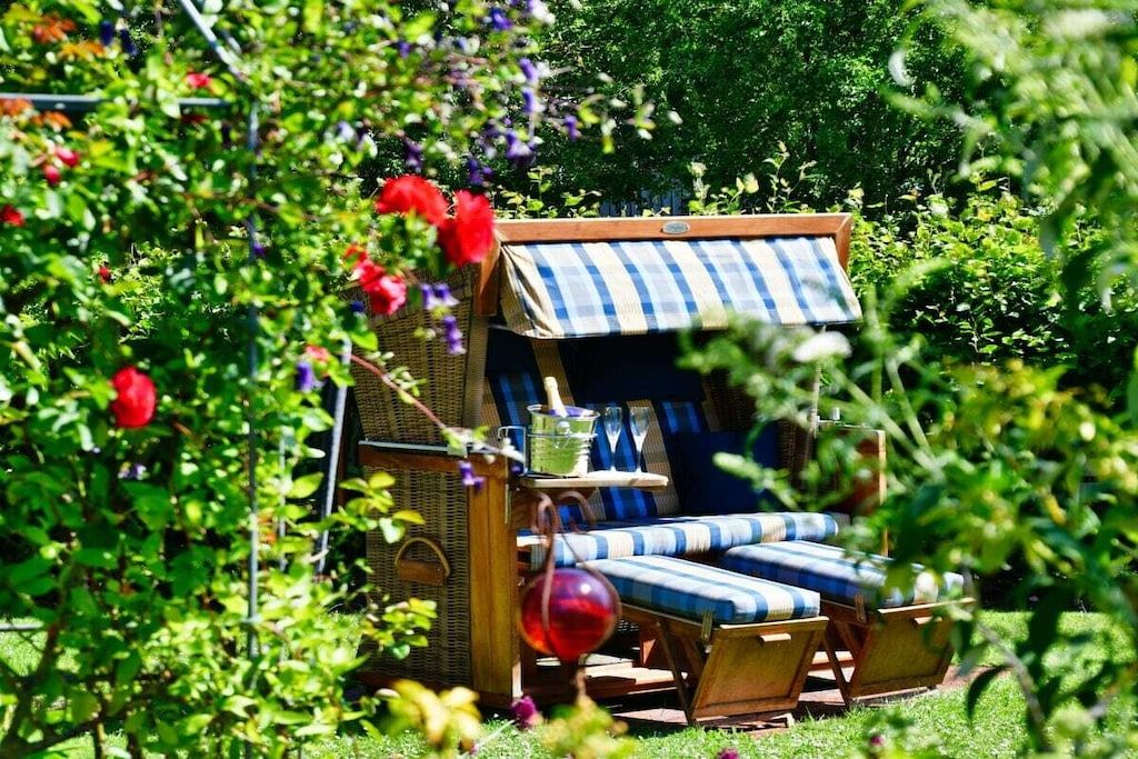 A garden setting with a small wooden structure featuring a checkered awning, a wicker chair, and a lounge chair with striped cushions.