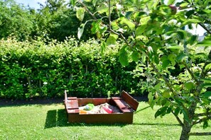 A wooden playbox with sand and toys on a grassy lawn, surrounded by a hedge and a tree with green leaves. - Holiday apartment with 2 bedrooms (Nordrhein-Westfalen - Kalkar)