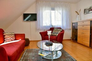 A living room with a red sofa, a glass coffee table, a television mounted on the wall, and a window with sheer curtains. - Holiday apartment with 2 bedrooms (Nordrhein-Westfalen - Kalkar)