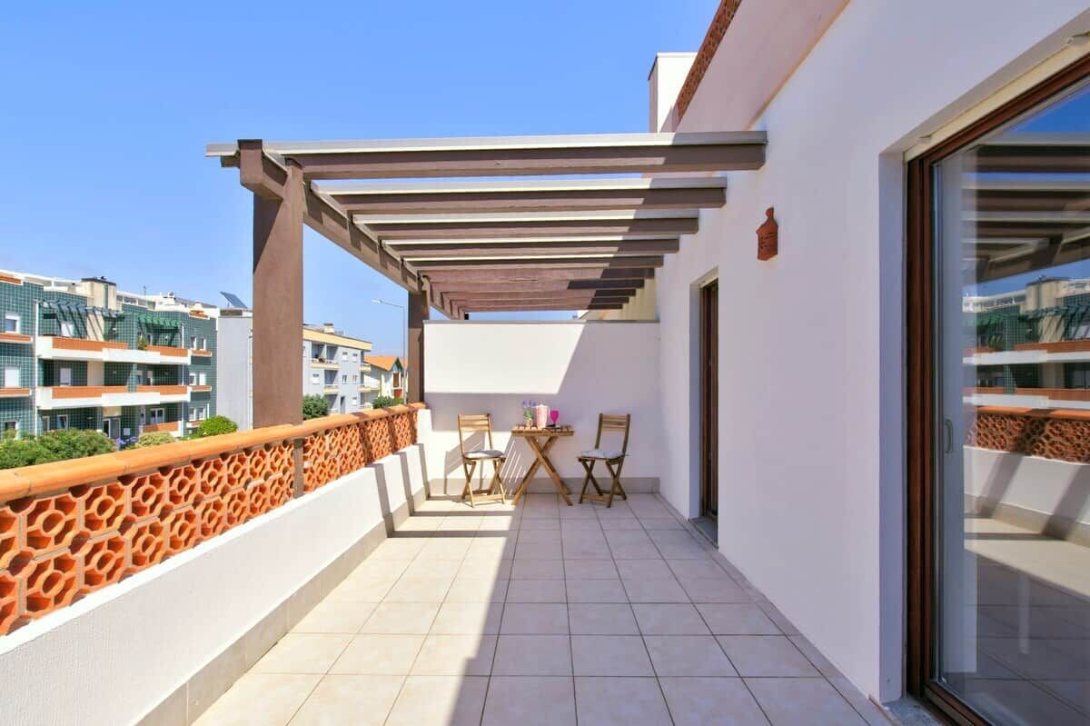 A balcony with a pergola, two chairs, a small table, and a view of neighboring buildings.