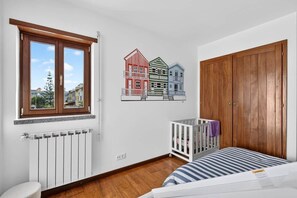 A bedroom with a window, a radiator, a crib, and a wooden wardrobe. - Lagosta Beach House (Gafanha da Boa Hora)
