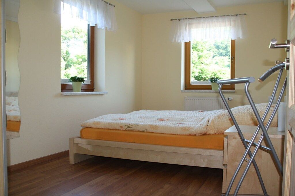 A single bed in a room with two windows, each with a curtain, a radiator, and a potted plant on the windowsill.