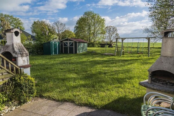 A grassy backyard with a stone barbecue grill, a swing set, and a small shed. - Holiday home with 1 bedroom (Boltenhagen OT Redewisch)