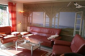 A room with a red leather sofa, a glass coffee table, and a view of the sea through a window with white shutters. - Holiday home with 1 bedroom (Boltenhagen OT Redewisch)