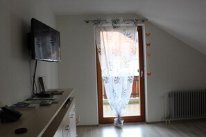 A room with a desk, a flat-screen TV mounted on the wall, a window with a curtain, and a radiator on the floor. - Holiday apartment with 1 bedroom (Endingen-Kiechlinsbergen)