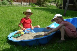Zwei Kinder spielen mit einem Spielzeug-Lkw und einer Schaufel in einem blauen Plastikbehälter auf der Wiese.