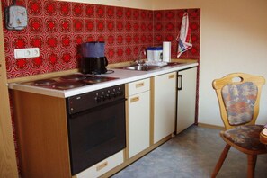 Une cuisine de taille réduite, équipée d’une cuisinière, d’un four et d’un évier. On y trouve une chaise à motifs floraux et une table en bois.