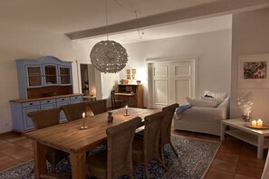 Un comedor con una mesa de madera y sillas de mimbre, una lámpara colgante y un sofá blanco al fondo.