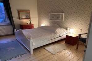 Een slaapkamer met een bed, nachtkastje, kapstok, spiegel en raam.