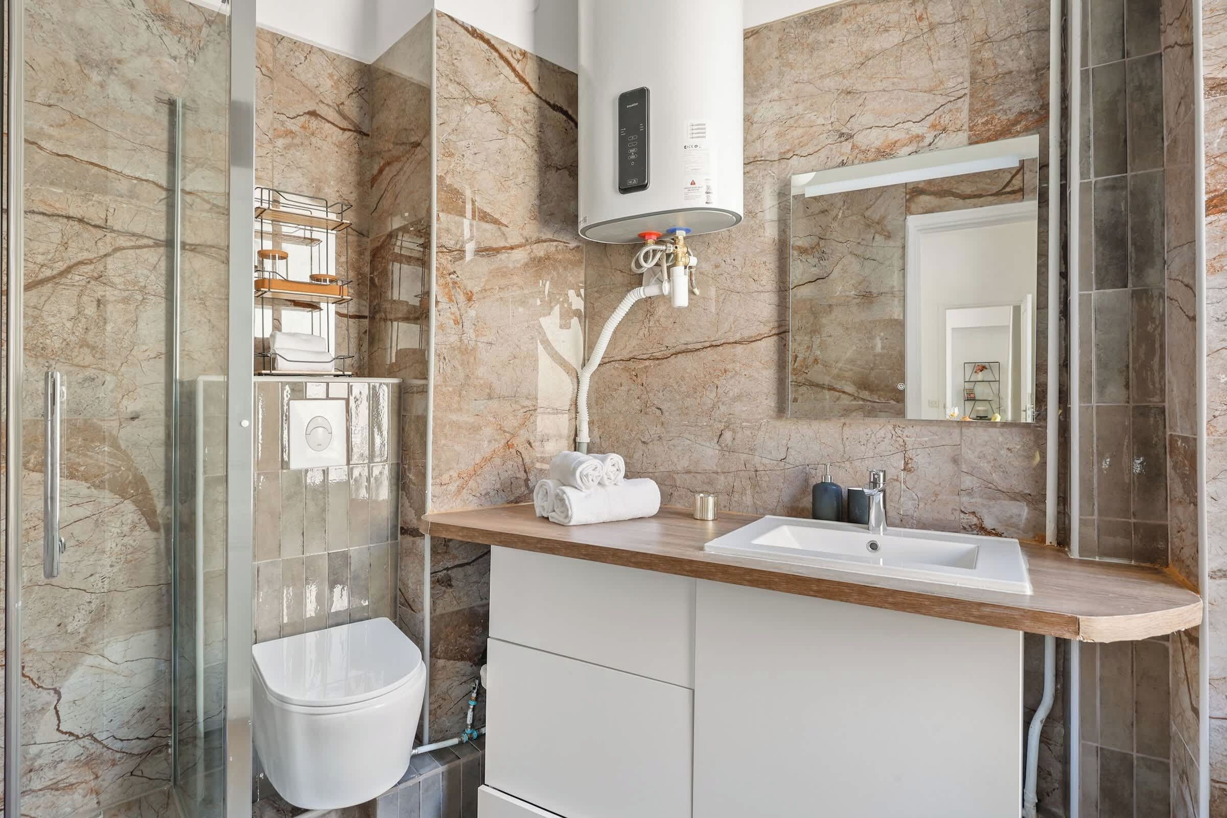 Een moderne badkamer met een marmeren spatwand, een glazen douchecabine, een witte wastafel en een toilet.