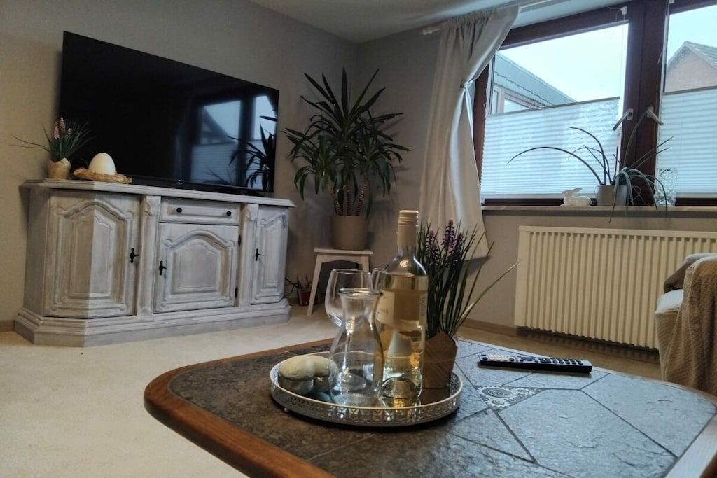 A living room with a flat-screen TV, a cabinet, a potted plant, a tray with glasses and bottles, and a remote control on a coffee table.