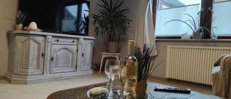 Een woonkamer met een flatscreen televisie, een kast, een potplant, een dienblad met glazen en flessen, en een afstandsbediening op een salontafel.