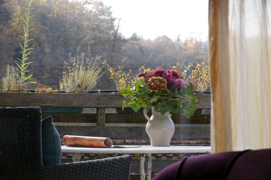 Eine Holzbank mit einer weißen Vase voller Blumen, einem eingerollten Gegenstand und einem Korbstuhl.