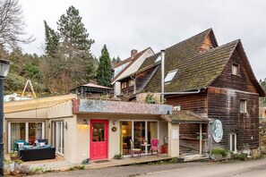 Un edificio rústico de madera con puerta roja, un pequeño porche con sillas y una ventana visible.