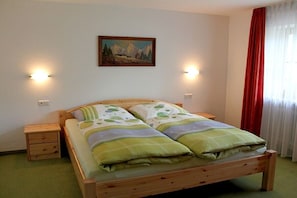 A single bed with a wooden frame, two bedside tables, a wall-mounted painting, and a window with red curtains. - Grünau on the ground floor (Schönau am Königssee)