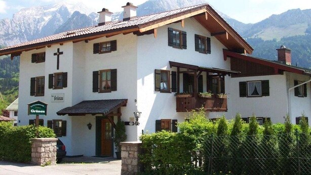 A two-story building with a cross on the facade, a sign reading 'Grünau,' and a mountain backdrop. - Grünau on the ground floor (Schönau am Königssee)