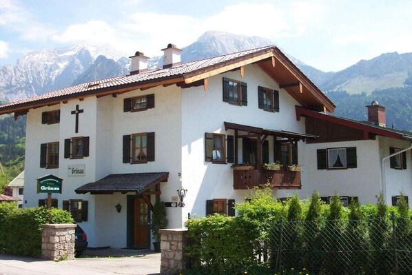 A two-story building with a cross on the facade, a sign reading 'Grünau,' and a mountain backdrop. - Grünau on the ground floor (Schönau am Königssee)